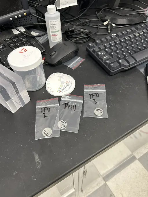 Three small bags on a lab bench, each holding a coin cell labeled with its formulation