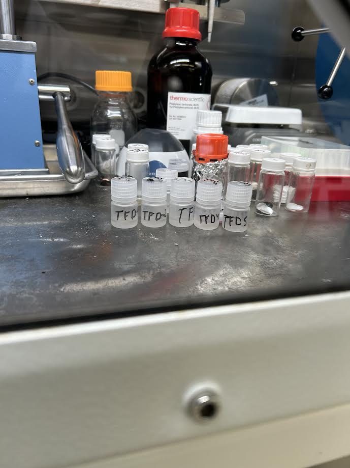 Small labeled vials of electrolyte formulations lined up inside a glovebox, seen through the glovebox window